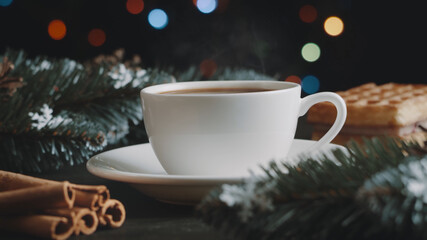 White cup of hot coffee on a brown wooden table. Green spruce branches, homemade waffles with souffle and cinnamon. Christmas lights and multicolored garland on background. New Year and Christmas