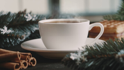 Obraz na płótnie Canvas White cup of hot coffee on a brown wooden table. Green spruce branches, homemade waffles with souffle and cinnamon. Winter morning. Window in the background. Cozy atmosphere. New Year and Christmas