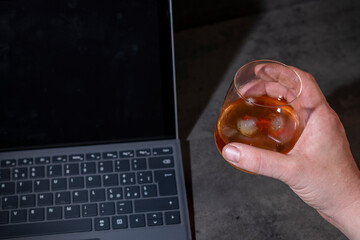 whiskey glass with ice in hand next to laptop on dark gray cement background