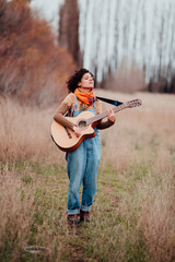 Young slim brunette woman playing guitar and singing in the field. Patagonian landscape background