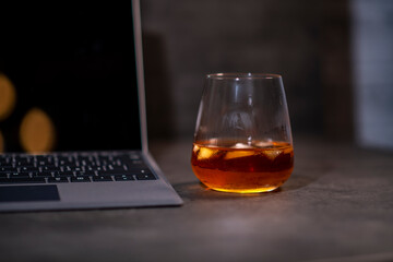 whiskey glass with ice in hand next to laptop on dark gray cement background