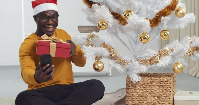 Afro american black man with glasses sits on floor of house near Christmas tree wears Santa Claus hat holds smartphone speaks to camera of mobile phone makes video for blog communicates in online chat