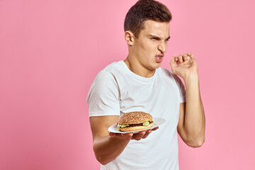 Emotional guy with hamburger on a plate and white t-shirt pink background cropped view of fast food calories