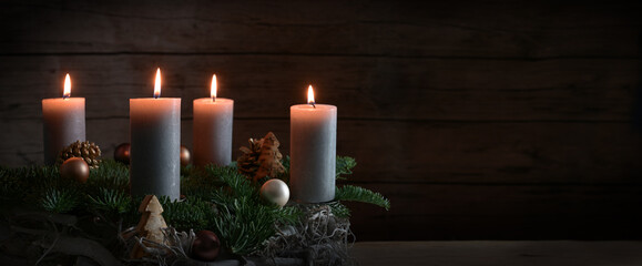 Four burning candles on an Advent wreath from fir branches with Christmas decoration against a dark rustic wooden wall, panoramic format with copy space, selected focus