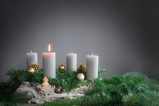 One of four candles is burning for the first Advent on fir branches with Christmas decoration against a grey background, copy space
