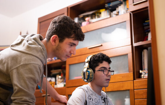 Staying at home: Two Caucasian brothers keep busy with their favorite video game. One is sitting with the gaming headset on his head and the other is standing next to him to suggest moves to take.
