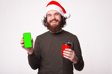 Handsome bearded man smiling and showing phone with green screen and holding cup of coffee to go