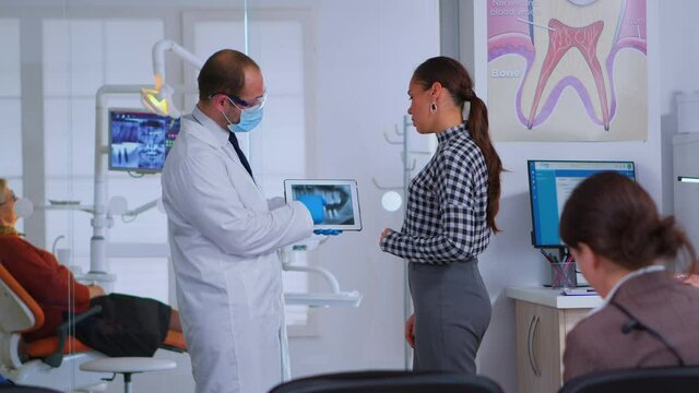 Dentist in waiting room of dental office talking with woman patient examining x-ray image on tablet while patients sitting on chairs in rception area. Doctor showing dental radiography, modern gadget