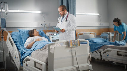 Obraz premium Hospital Ward: Friendly Latin Doctor Talks to Beautiful Female Patient Resting in Bed, Uses Tablet Computer, Explains Test Results. Physician Talks to Woman Recovering after Successful Surgery