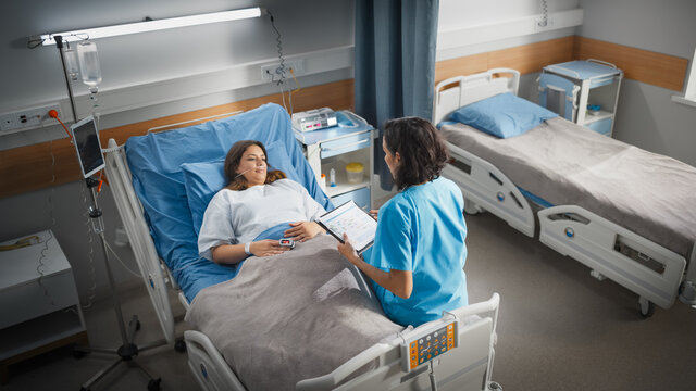 Hospital Ward: Friendly Hispanic Head Nurse Fills Medical History Form, Talks to Female Patient Recovering in Bed. Professional Nurse Helps Happy Woman Get Better after Surgery. High Angle Shot - Powered by Adobe