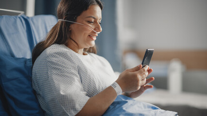 Medical Hospital Ward: Portrait of Beautiful Latin Woman with Nasal Cannula Resting in Bed, Uses Smartphone to Video Chat Conference with Her Family, Friends. Recovering after Successful Surgery