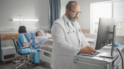 Hospital Ward: Professional Experienced Latin Doctor / Surgeon Uses Medical Personal Computer. In the Background Head Nurse Sitting with a Patient Recovering After Surgery in Bed 