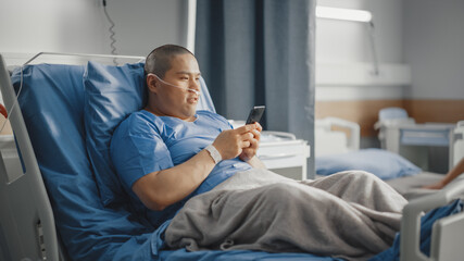 Obraz premium Medical Hospital Ward: Portrait of Handsome Chinese Man with Nasal Cannula Resting in Bed, Uses Smartphone, Playing Video Games, Doing e-Shopping, Chatting with Friends. Surgery Recovery Room.