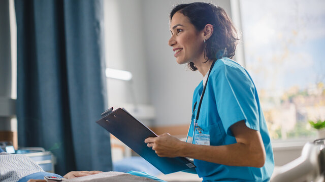 Hospital Ward: Portrait Of Friendly Hispanic Head Nurse Fills Medical History Form, Talks To Anonymous Patient Recovering In Bed. Doctor Helps Person Get Better. Close-up Portrait Shot.