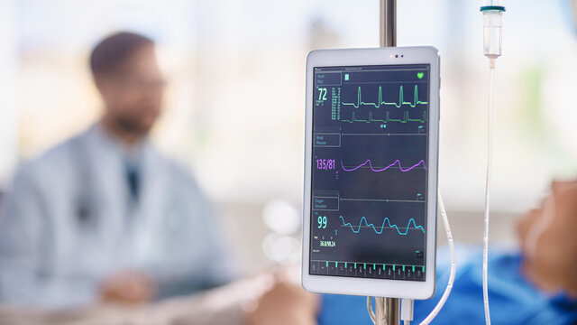 Medical Hospital Ward: Close-up Shot of a Digital Tablet Mounted near Dropper Showing Medical Software with Patient's Herat Rate, Blood Pressure numbers. In Background Blurred Docktor Helping Patient