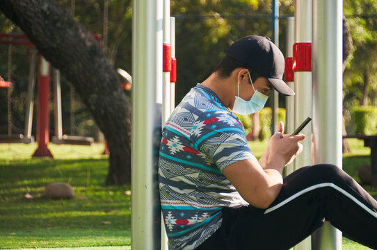 Young Man Using His Cell Phone Wearing A Medical Mask After His Exercise Routine In The Park 