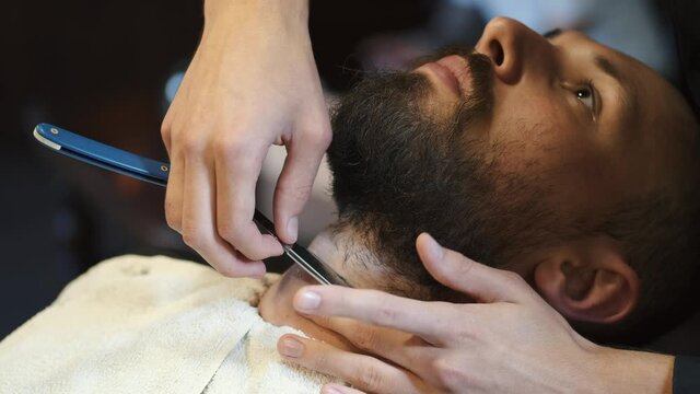 Unrecognizable barber shaving a customer with a classic razor. Man and barber with straight razor shaving beard, slow mo.