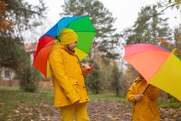 Cute happy little boy and a girl - brother and sister - in identical yellow costumes and hats walking in the forest with rainbow-colored umbrellas. Cosiness, family