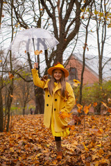 cute beautiful teenage brunette girl in an orange hat, dress and coat holding a transparent umbrella with falling leafes over her. Cosiness, autumn