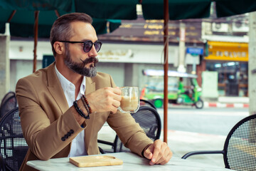 Confidence Caucasian businessman in casual suit sit on outdoor chair outside coffee shop with drinking cafe latte coffee on the table. Handsome beard man relax and enjoy holiday vacation in the city.