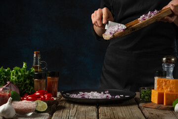 Professional chef in black uniform pours chopped onion on the plate. Backstage of cooking traditional mexican tacos on rustic wooden table with ingredients background. Cooking process.