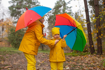 Cute happy little boy and a girl - brother and sister - in identical yellow costumes and hats walking in the forest with rainbow-colored umbrellas. Cosiness, family