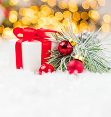 Present with red ribbon, pine or spruce branch with red balls on the snow on the background of blurred Christmas tree and lights of garland.