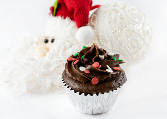 Close up view of a Christmas chocolate cupcake with red, green and white sprinkles.  Santa and lace ball ornament blurred in background.  White background.