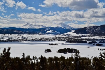 Snow covered Rocky Mountain peaks surrounded by white puffy clouds and a snow covered lake near Vail, Colorado  - obrazy, fototapety, plakaty