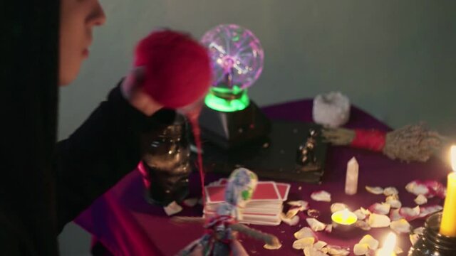 Dark magic voodoo witch performing ritual and casting spell piercing doll with needles sitting in dark room with burning candles.