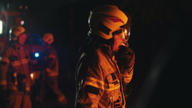 Close up of firefighter using radio to speak with his collegues. Fire engine with emergency lights and two more firemen in the background. High quality 4k footage