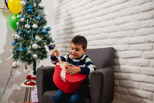 A delighted 5-year-old boy opens his Christmas present with a ribbon and sits in a house on a chair next to the New Year's tree and balloons. Happy New Year and Xmas