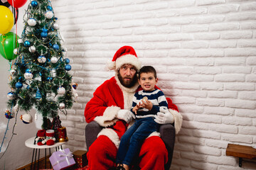 A cute 5-year-old boy is sitting in Santa's lap in the house next to a decorated Christmas tree. Happy New Year and Merry Christmas