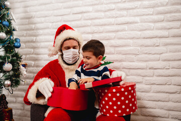 A smiling 5 year old boy sitting in Santa's lap with a face mask and watching a xmas gift next to a Christmas tree. New Year and Christmas pandemic COVID -19 coronavirus