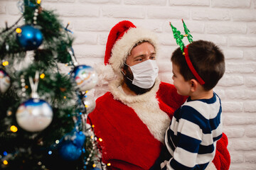 A cute smiling boy in the arms of Santa Claus with a face mask for Christmas next to the xmas tree. Happy New Year and merry xmas during the COVID -19 CORONAVIRUS pandemic