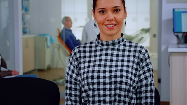 Portrait of smiling woman patient looking on webcam sitting on chair in waiting room of stomatological clinic. Stomatologist assistant typing on pc in dental office while doctor working in background.