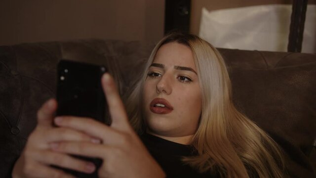 Young blond beautiful woman stretching on the sofa is playing game with smartphone and feeling sad