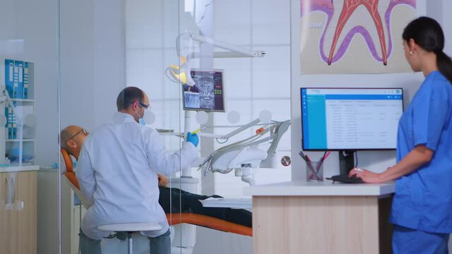 Patients asking for help filling in dental registration form preparing for exemination. Senior woman sitting on chair in waiting area of crowded orthodontist office while doctor working in background