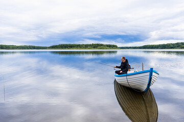 Fototapeta na wymiar An elderly retired man is fishing from a wooden boat on the lake with calm water