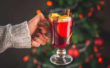 A woman's hand in a warm sweater is holding a cup of aromatic hot mulled wine against the...