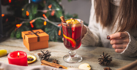 Woman in a warm sweater is drinking aromatic hot mulled wine on the background of a Christmas tree with lights. Concept of a holiday atmosphere and cozy winter mood