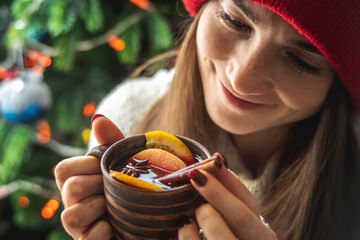 Woman in a warm sweater is holding a cup of aromatic hot mulled wine on the background of a Christmas tree with lights. Concept of a holiday atmosphere and cozy winter mood