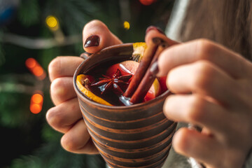 A woman's hand is holding a cup of aromatic hot mulled wine against the background of a Christmas tree with lights. Concept of a festive atmosphere