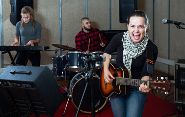 Fototapeta na wymiar Portrait of active excited cheeful girl rock singer with guitar during rehearsal with male drummer and female keyboardist in studio