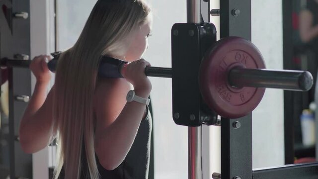Back view of blonde woman does squats with barbell on smith machine at gym
