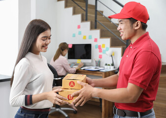 Delivery personnel in red uniform delivering packages of goods to home with a smile and happy face. Young Asian business receiving boxes from the postman at the home office. Selective focus on hand