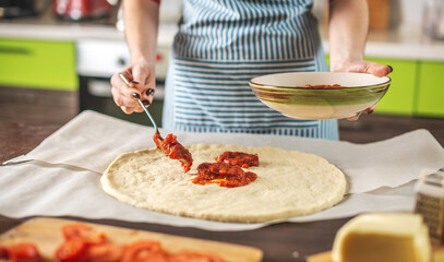 Female chef in an apron is putting tomato sauce on a raw pizza. Cooking delicious pizza at home in the kitchen.