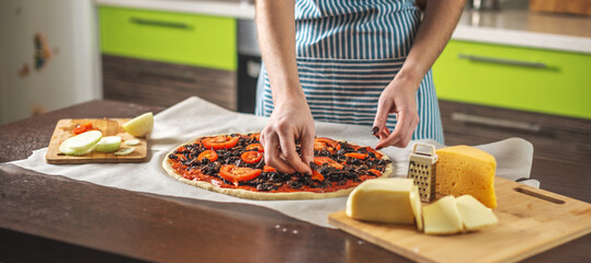 Female chef in an apron is putting the tomatoes on a raw pizza. Cooking delicious pizza at home in the kitchen