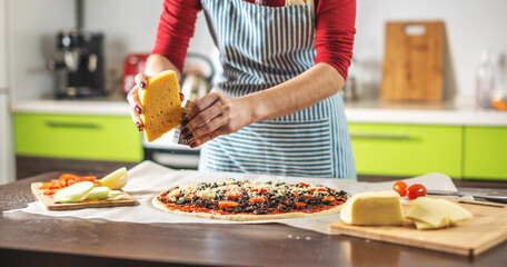 Female chef in an apron is rubbing cheese on a raw pizza. Cooking delicious pizza at home in the kitchen.