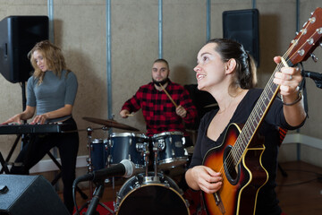 Fototapeta na wymiar Rehearsal of music band. Positive female guitar player and singer practicing with band members in recording studio